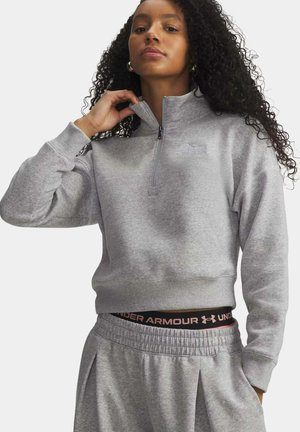 Vrouw met lang krullend haar die een grijze trui met een kwart ritssluiting en bijpassende joggingbroek draagt, en haar kraag met één hand aanpast.