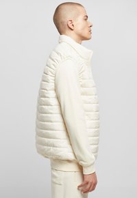 Kreemikas värvi puffer-vest horisontaalse õmblusmustriga, kõrge krae ja tõmblukuga. Kantakse koos sobiva ümarakaelusega dressipluusi ja joggeritega.