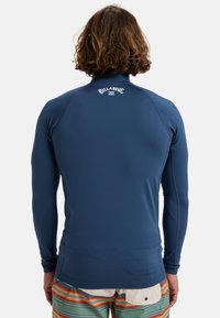 Marinblå långärmad rash guard tillverkad av stretchigt material, med platta sömmar och en logotyp på baksidan. Matchas med randiga shorts.