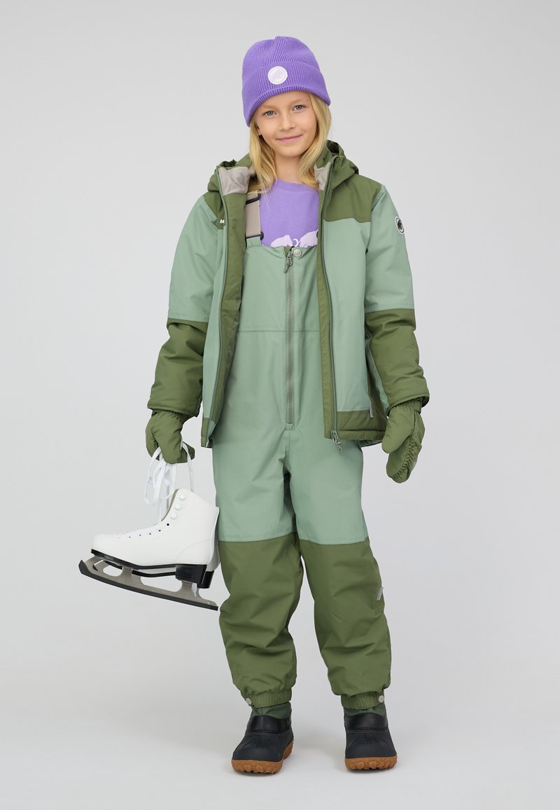 Bambino in tuta invernale verde e cappello viola che tiene i pattini da ghiaccio bianchi, in piedi davanti a uno sfondo uniforme.