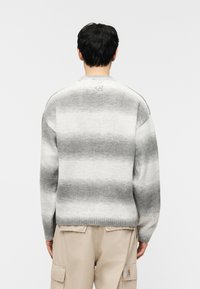 Άτομο που φοράει γκρι λευκό ριγέ πουλόβερ, με την πλάτη γυρισμένη, σε συνδυασμό με μπεζ παντελόνι cargo, σε λευκό απλό φόντο.