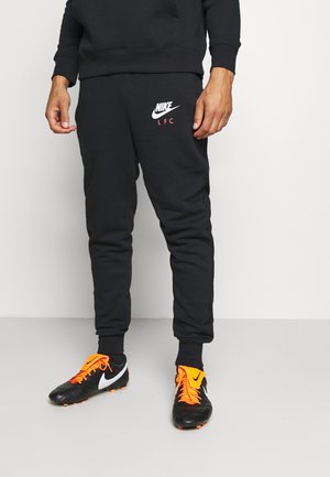 Personne portant un pantalon de survêtement Nike LFC noir et des chaussures de football noires avec des lacets et détails orange vif, debout devant un fond blanc.