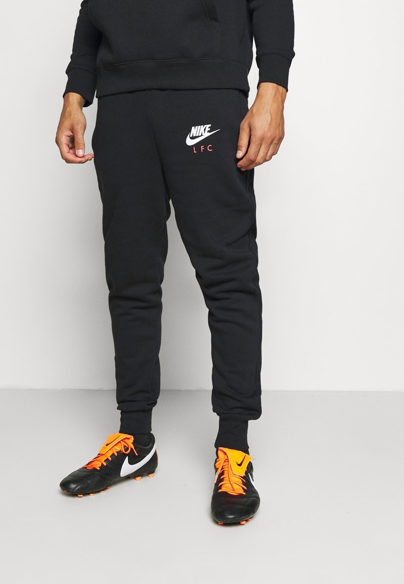 Personne portant un pantalon de survêtement Nike LFC noir et des chaussures de football noires avec des lacets et détails orange vif, debout devant un fond blanc.