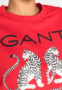 Rød bomulds T-shirt med grafisk tryk af to hvide tigre med sorte striber, ledsaget af mærkenavnet "GANT" i fede sorte bogstaver.