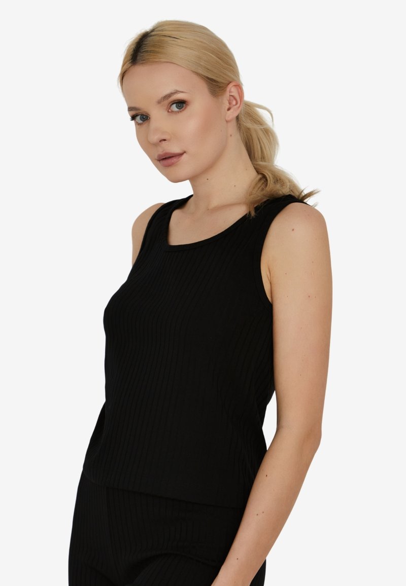 Blondynka z kucykiem, ubrana w bezrękawnik w czarny prążkowany wzór i pasujące czarne spodnie, skierowana lekko w lewo z neutralnym wyrazem twarzy.