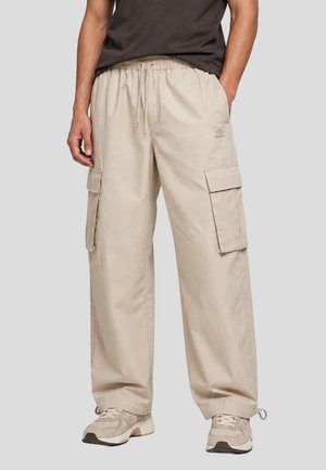 Uomo che indossa pantaloni cargo beige con coulisse in vita e sneakers beige, in piedi con una mano in tasca su sfondo neutro.