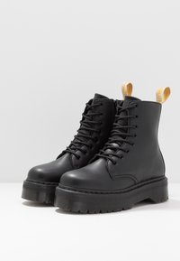Stivali da combattimento in pelle nera con suola a piattaforma spessa, punta rotonda e design con lacci. Presentano anelli gialli a forma di occhielli sul retro.