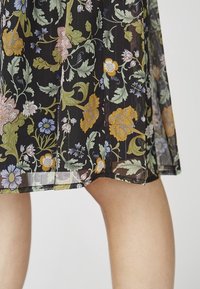 Bloemjurk van doorzichtig zwart materiaal, met meerkleurige bloempatronen in geel, roze en blauw. De zoom is licht gebogen.