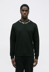 Moški nosi črno dolgouhanico s sweatshirtom, na katerem je besedilo "VERSACE JEANS COUTURE" okoli izreza, in stoji pred enobarvno ozadje.