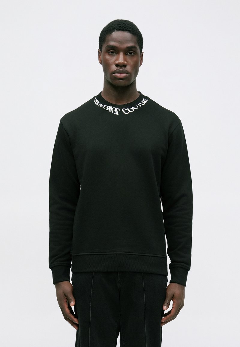 Moški nosi črno dolgouhanico s sweatshirtom, na katerem je besedilo "VERSACE JEANS COUTURE" okoli izreza, in stoji pred enobarvno ozadje.