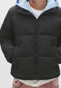 Sort dunjakke med blå hætte, lynlåslukning foran og quiltet mønster. Har et lille logo på brystet og to sidelommer.