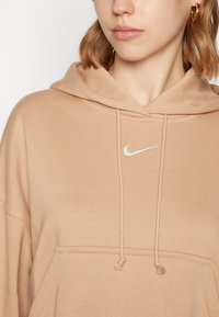 Hoodie bege com um capuz ajustável, bolso frontal e um logo prateado da Nike no peito; feito de tecido suave com uma textura lisa.