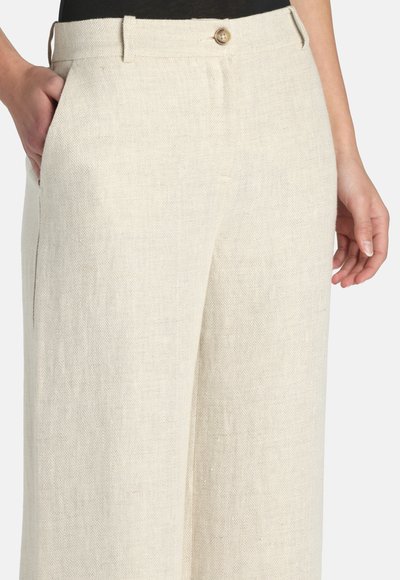 Pantalon beige taille haute à jambes larges avec fermeture à boutons et passants pour ceinture, porté avec une main dans la poche et l'autre détendue sur le côté.