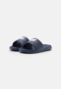 Tengerészkék műanyag strandpapucs széles pánttal, amelyen fehér logó található, valamint texturált talpbetéttel a tapadás érdekében.