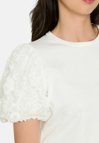 Balta blūze ar teksturētām uzpūstām piedurknēm, kas izgatavotas no mežģīnes ar ziedu rakstiem, gludu korpusa audumu un apaļu kakla izgriezumu.