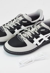 Paire de baskets Asics noires et grises avec des bandes blanches et des lacets, accompagnée d'une paire supplémentaire de lacets blancs sur fond blanc.
