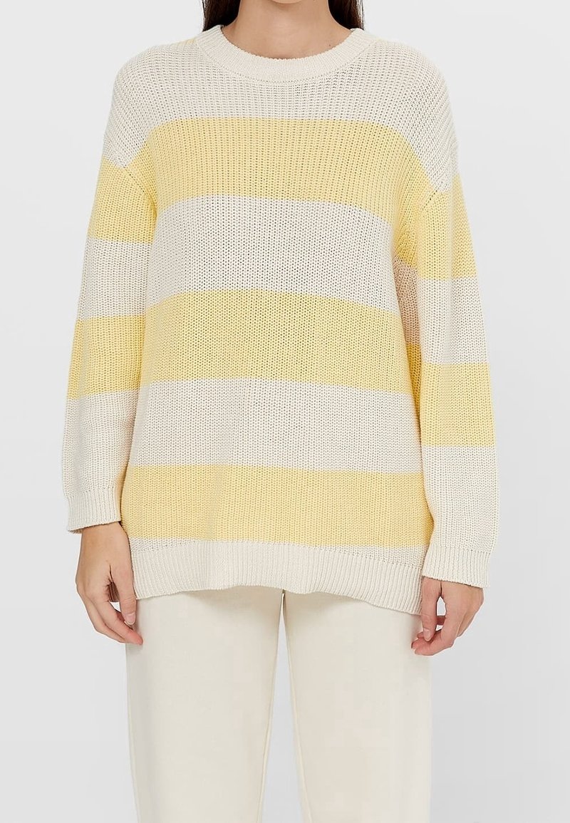 Grovere gebreide trui met brede horizontale strepen in lichtgeel en wit, gedragen met lichtgekleurde broek.