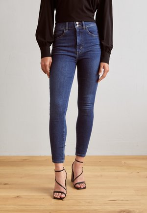 Vrouw draagt donkerblauwe skinny jeans met hoge taille en zwarte hoge hakken met bandjes, staand op een houten vloer.