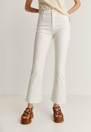 Witte high-waisted jeans met een flared leg ontwerp, voorzien van voorzakken en gestikte details. Gekoppeld aan bruine gespsandalen met platform.