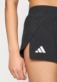 Shorts de sport noirs avec une fente latérale, une ceinture élastique et un logo Adidas blanc sur l'ourlet, fabriqués en tissu léger et respirant.