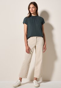 Tyrkysové tričko s krátkým rukávem a nařaseným výstřihem, v kombinaci s béžovými kalhotami s širokými nohavicemi. Model má na sobě bílé tenisky, jednoduchý design, měkký materiál.