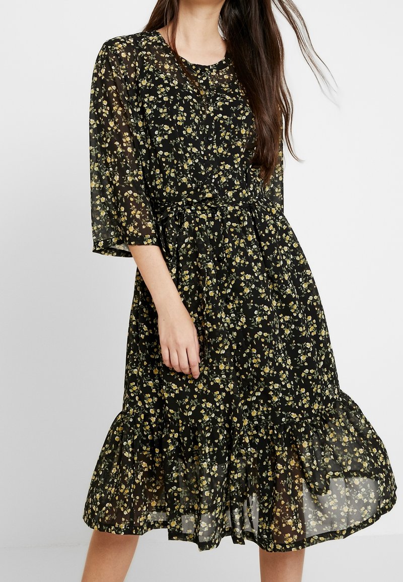 Robe noire à fleurs avec des fleurs jaunes, tissu transparent, encolure ronde, manches courtes, taille cintrée et ourlet froncé.