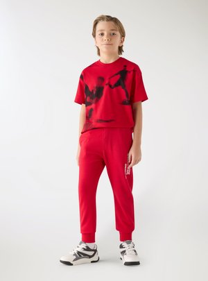 Ragazzo in piedi con maglietta rossa con grafiche nere di calciatore, pantaloni della tuta rossi e scarpe da ginnastica bianche e nere su sfondo bianco.