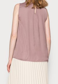 Bezramenná mauve blůza s detailem klíčové dírky na zádech, vyrobená z lehkého materiálu, měkce splývá přes plisovanou béžovou sukni.
