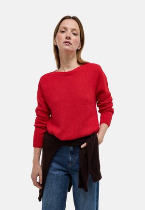 Femme portant un pull en maille rouge, un jean bleu et un pull foncé noué autour de la taille, debout devant un fond blanc uni.