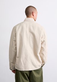 Beige cotton jacket with a classic collar, visible stitching, and buttoned cuffs, worn over olive green pants. Back view.