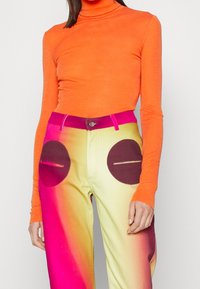Kvinna iklädd en ljus orange långärmad polotröja och högmidjade byxor med rosa, gula gradienter och mörka, cirkulära ficklappar.
