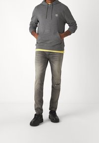 Gray hoodie with drawstring, logo patch; layered over a yellow shirt. Lightly distressed gray jeans; black sneakers with textured surface.