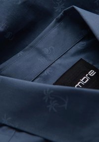 Camisa azul marino con una textura suave, que presenta un sutil patrón de flamencos y palmeras. La etiqueta está cosida en tela negra.