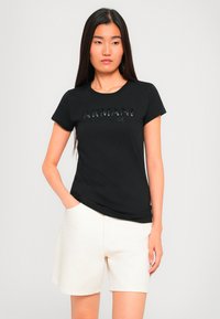 Kvinne med langt mørkt hår iført en svart Armani t-skjorte og hvite shorts, stående med den ene hånden i lommen mot en ensfarget bakgrunn.