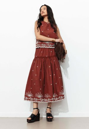 Femme debout contre un mur blanc, portant un haut sans manches couleur rouille et une jupe assortie avec broderie blanche, tenant un sac marron à franges.