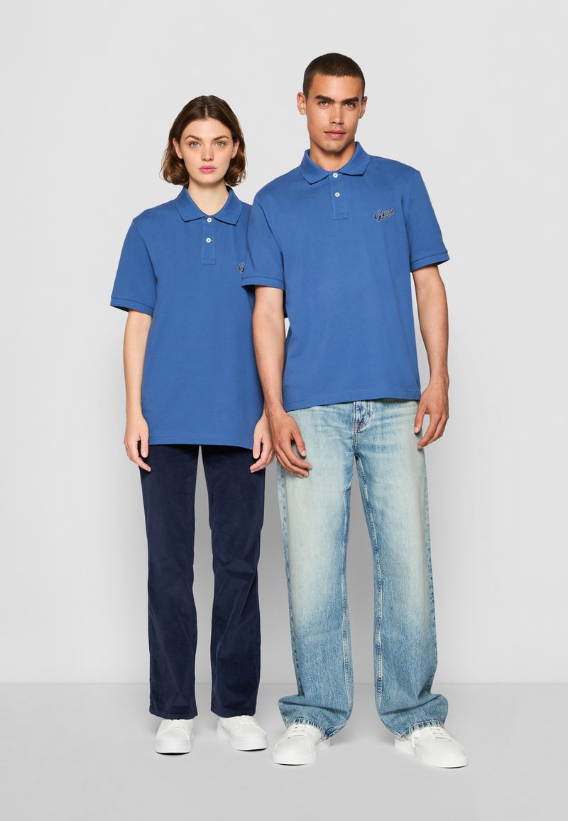 Chemises polo bleues à manches courtes, en coton, avec un petit logo sur la poitrine gauche. Associées à des pantalons en denim foncé et clair.