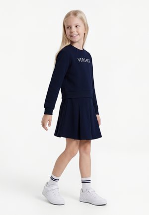 Młoda dziewczyna z długimi blond włosami, ubrana w granatową bluzę Versace i plisowaną spódnicę, białe trampki oraz paskowane skarpetki, stoi i się uśmiecha.