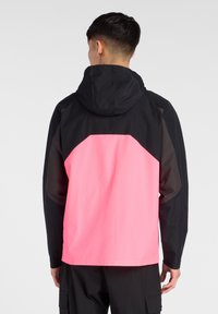 Veste imperméable avec une capuche et des manches noires, et un panneau arrière rose vif. Texture lisse, design sportif et coupe pratique.