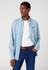 Lys blå skjorte med knaplukning lavet af denim, med en lomme, parret med en hvid t-shirt og mørkeblå jeans, sikret med et brunt bælte.