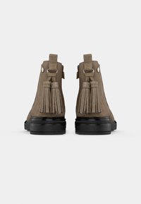 Bottines en daim marron avec des semelles noires, vues de l'arrière, chacune présentant un détail de boucle et de frange.