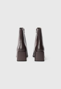 Bottines en cuir brun avec un talon carré, présentant une fermeture éclair et une texture lisse et polie. Le design est élégant et moderne.