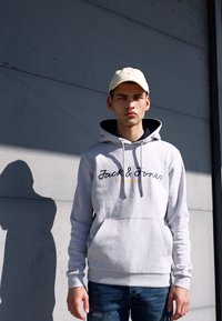 Grå bomulds hættetrøje med en frontlomme, snore og logo tekst. Båret med blå jeans og en beige kasket. Baggrunden er en tekstureret væg.