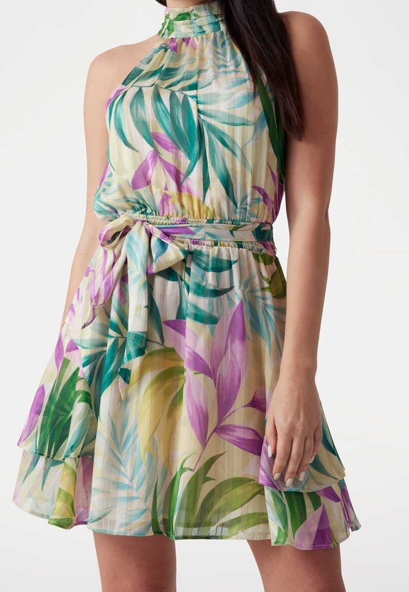 Robe sans manches à col halter avec imprimé multicolore de feuilles tropicales, taille cintrée avec lien en tissu et jupe superposée portée par une femme aux cheveux foncés.