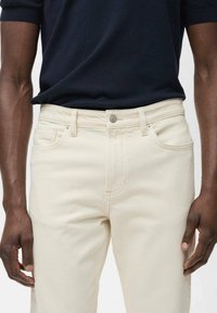 Blugi bej din denim cu un design ajustat și o cusătură subtilă. Prezintă un închidere cu buton în față și buzunare laterale. Materialul are o textură netedă.