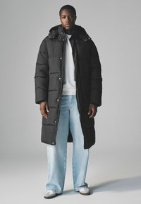 Sort pufferjakke med hætte, der har kviltede sektioner, trykknaplukninger og et langt design. Bæres over en hvid sweatshirt og lyseblå jeans.