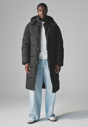 Sort pufferjakke med hætte, der har kviltede sektioner, trykknaplukninger og et langt design. Bæres over en hvid sweatshirt og lyseblå jeans.