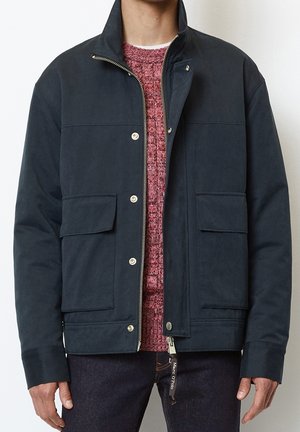 Homme portant une veste bleu marine foncé zippée et à boutons pression avec poches à rabats, par-dessus un pull en maille rouge et un pantalon sombre, debout devant un fond blanc.