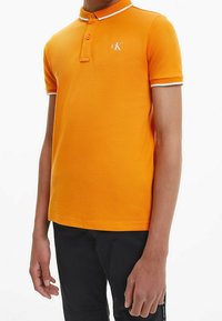 Oranžové polo tričko se strukturovaným povrchem, s límečkem, dvěma knoflíky a bílými akcenty na rukávech. Logo je umístěno na hrudi.