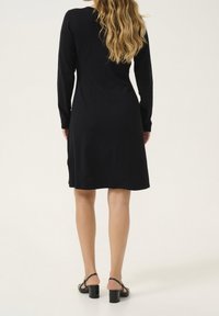 Robe noire à manches longues avec une coupe ample, une longueur jusqu'aux genoux et une texture lisse. Portée avec des sandales à talons carrés noires.