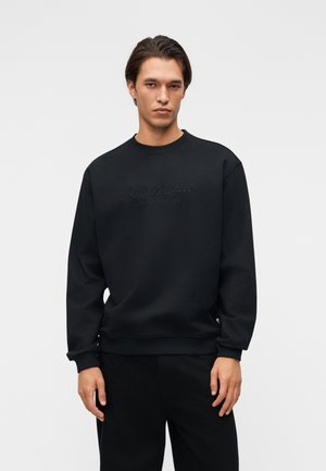 Young man with dark hair wearing a black long-sleeve sweatshirt with subtle "Carlo Colucci" text, standing against a light gray background.